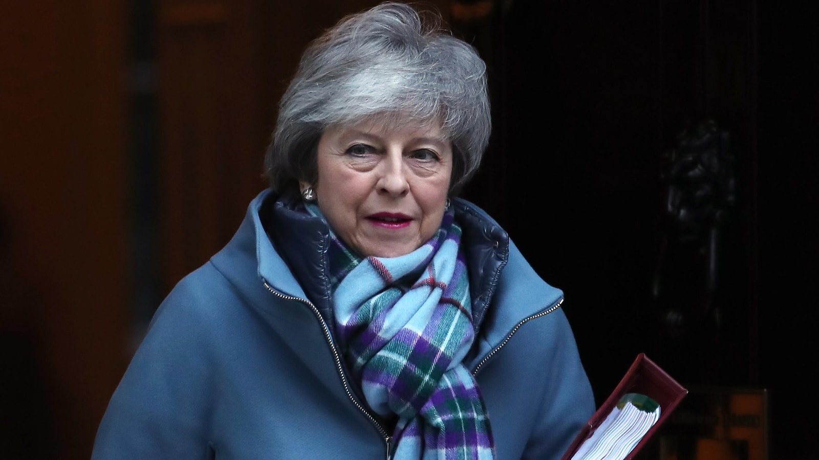 Theresa May, vêtue d'un manteau et d'un foulard, sort du 10 Downing Street avec un porte-document sous le bras.