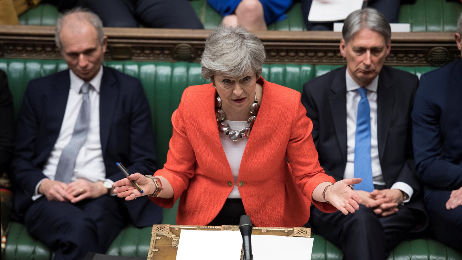 Theresa May debout à la Chambre des communes, parle au podium, devant un micro, alors qu'on voit des députés derrière elle.