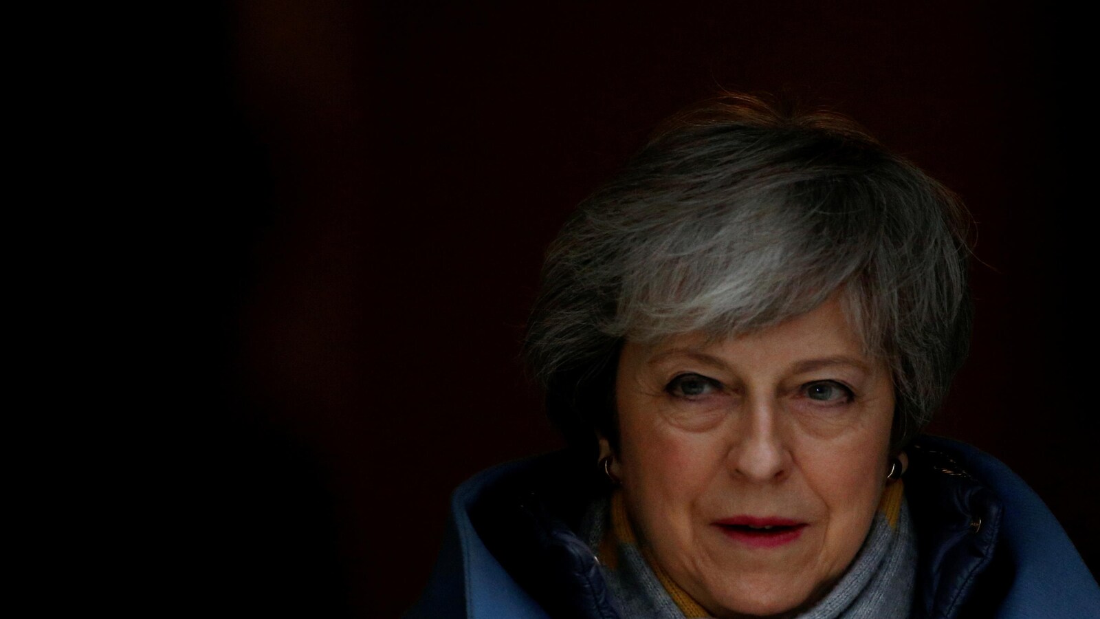 Dans l'ombre, le visage de Theresa May apparaît. Ses yeux verts brillent, ses cheveux sont courts et gris, elle arbore une frange.