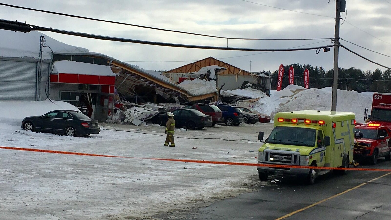 Derrière un périmètre de sécurité, une partie du toit d'un bâtiment est affaissée sur des véhicules stationnés. Une ambulance est sur les lieux.