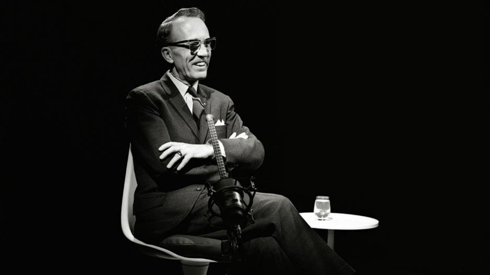 Une photo en noir et blanc d'un homme dans la cinquantaine souriant assis sur une chaise.