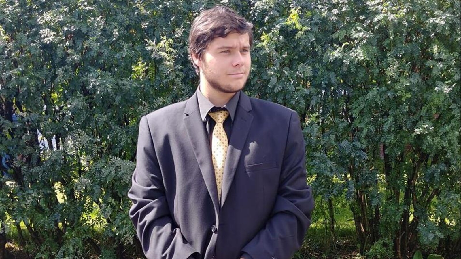 Un jeune homme porte un veston bleu et une cravate jaune. Il pose devant la caméra.