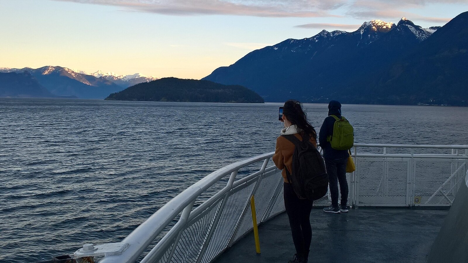 Deux passagers observent debout des montagnes enneigées depuis le pont d'un traversier qui quitte Horseshoe Bay en Colombie-Britannique.