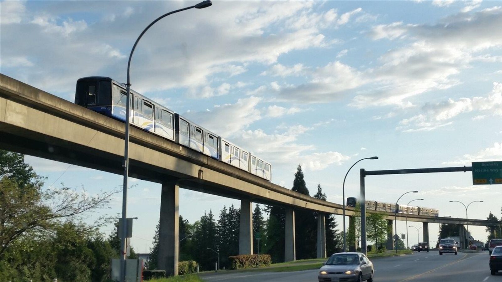 Deux trains du réseau de Skytrain de TransLink circulent au-dessus d'une rue sur laquelle passent des voitures.