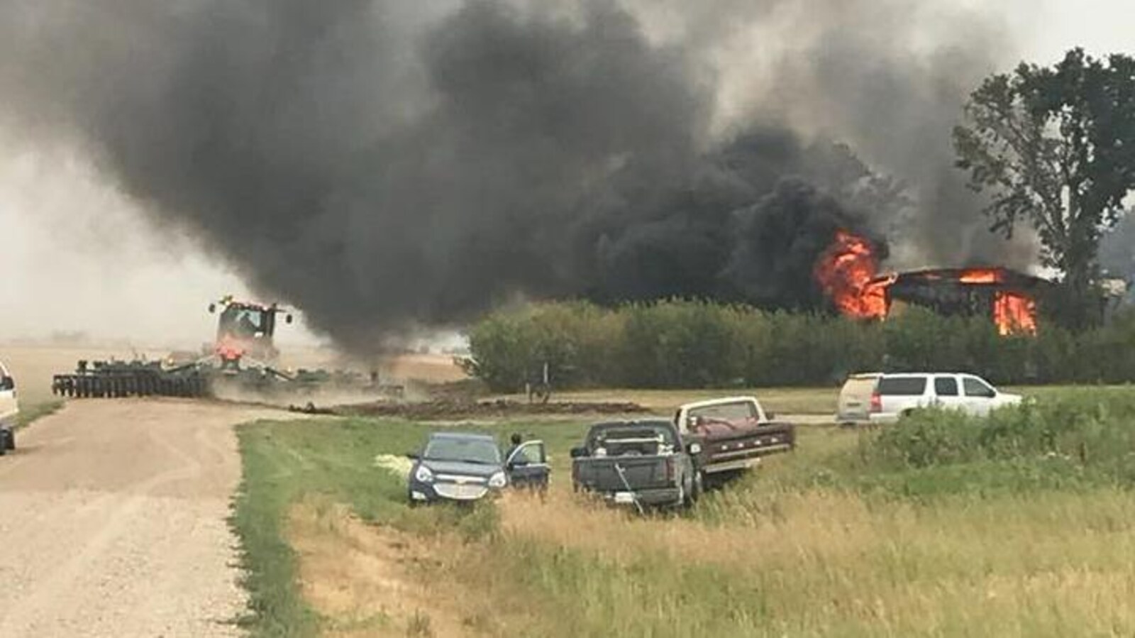 Une épaisse fumée s'élève au-dessus d'une maison en flammes. Plusieurs voitures sont stationnées derrière les arbres qui bordent la maison. À gauche, un homme dans un tracteur s'assure que les flammes ne se dispersent pas dans les champs.
