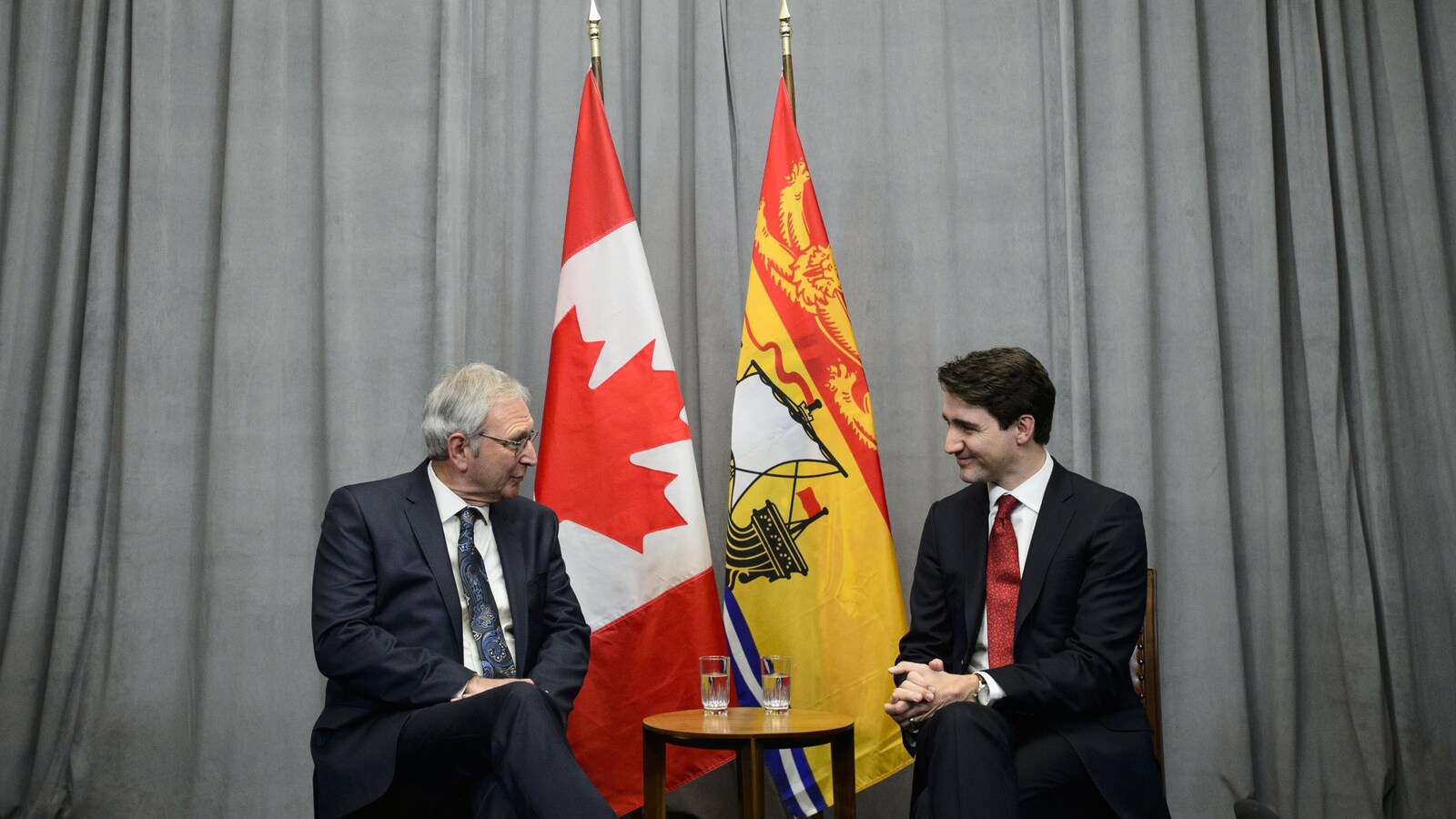 Blaine Higgs, à gauche, et Justin Trudeau, à droite, devant un rideau gris et deux drapeaux, l'un du Canada et l'autre du Nouveau-Brunswick.