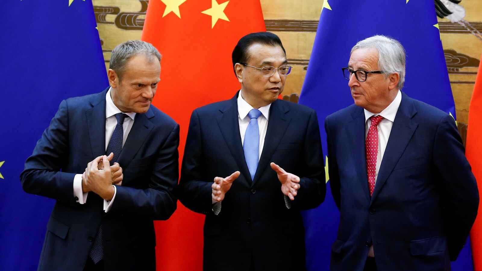 Trois hommes en veston-cravate, le 16 juillet 2018, se tiennent côte à côte devant des drapeaux étoilés et se parlent entre eux. Un des hommes est asiatique.