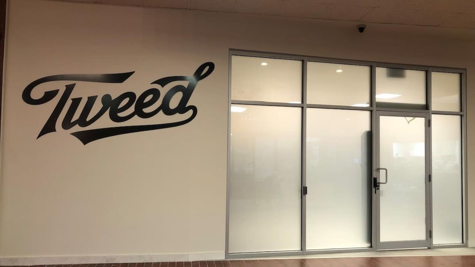 Sur un mur blanc, il est écrit Tweed en grand et en noir à côté d'une porte et de fenêtres en verre givré.