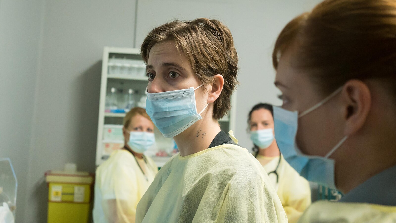 Entourée de plusieurs personnes avec des masques chirurgicaux, la comédienne, porte aussi un masque et une blouse d'hôpital.