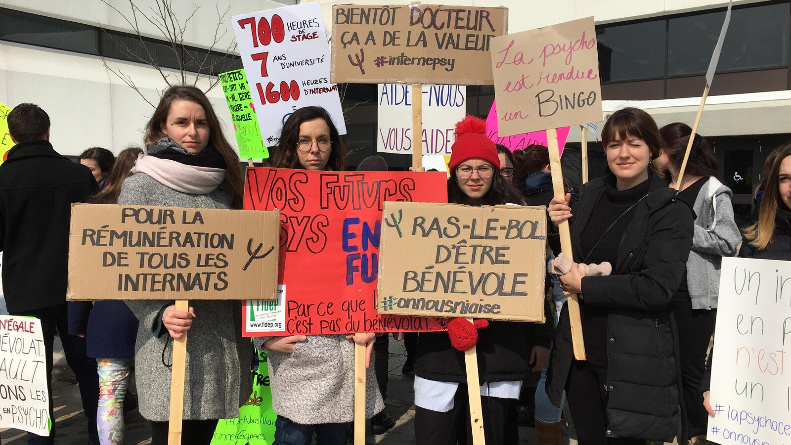 Quatre étudiantes tiennent des pancartes où des slogans sont inscrits. Il peut y lire «Pour la rémunération de tous les internats» et «La psycho est rendue un bingo»