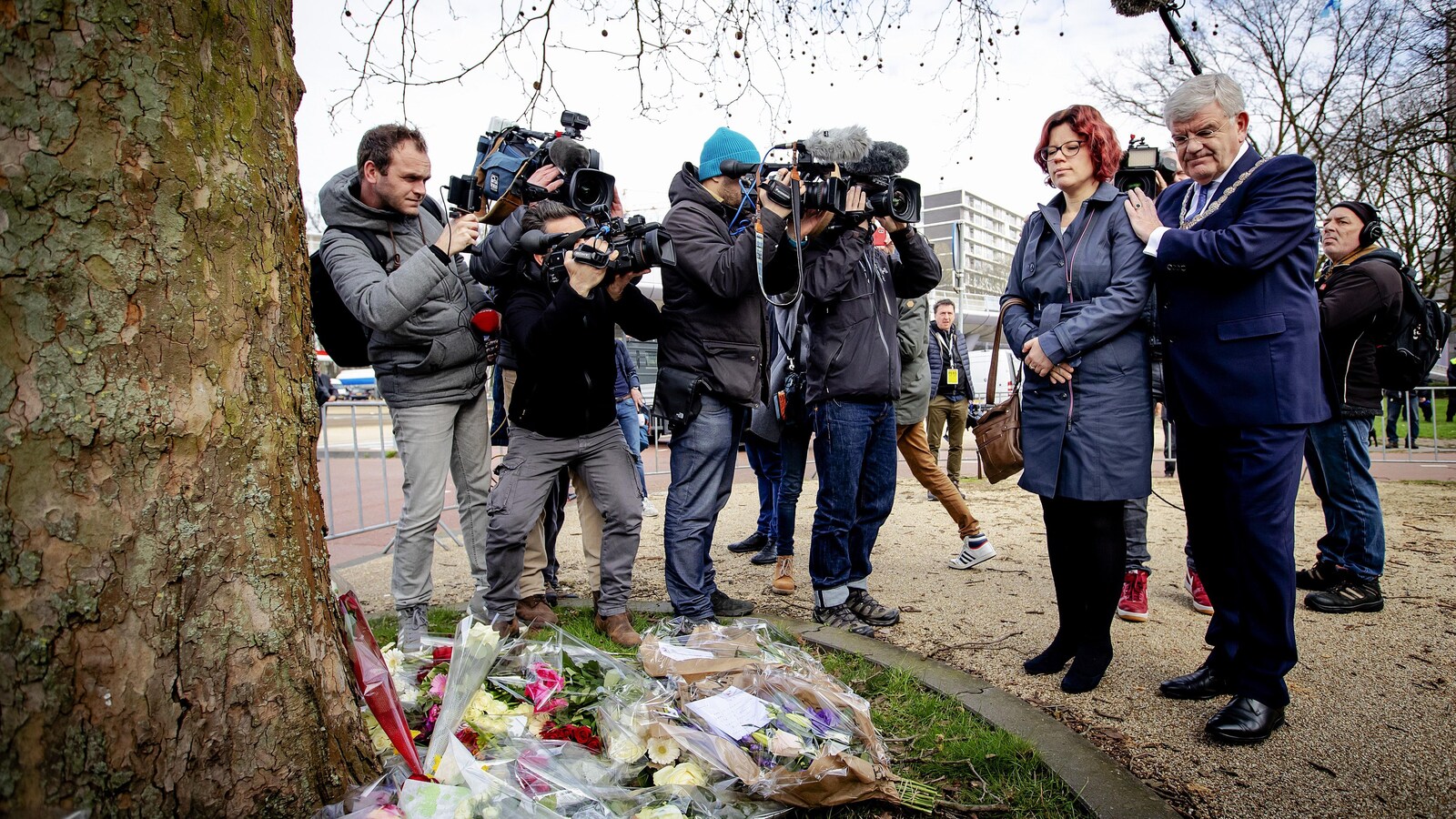 À gauche de l'image, des cameramen filment le maire, aux cheveux blancs et à la mine contrite et une conseillère municipale aux cheveux rouges, qui se recueillent devant un arbre. À terre, des fleurs sont déposées.