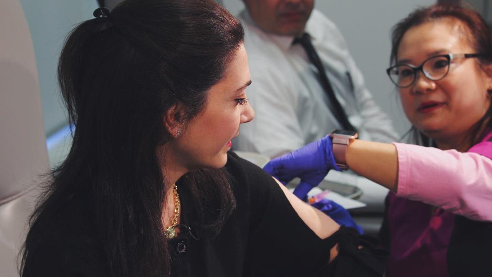 Une femme reçoit une piqûre, donnée par une autre femme avec des gants de la latex. Un homme, assis derrière, observe la vaccination.
