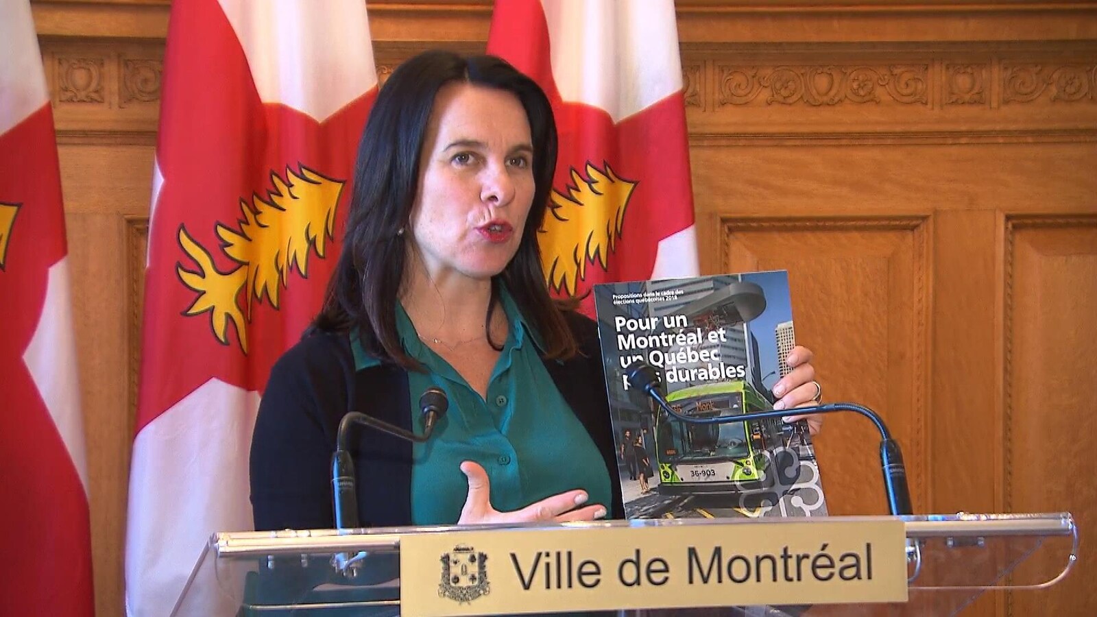 Une femme tenant un livret sur lequel on peut lire « Pour un Montréal et un Québec plus durables » parle derrière un lutrin. Cette femme se trouve devant des drapeaux.