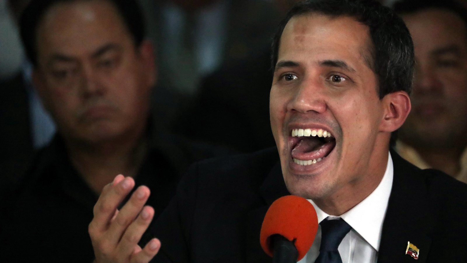 Juan Guaido a la bouche ouverte devant un micro orange avec des hommes en arrière-plan.