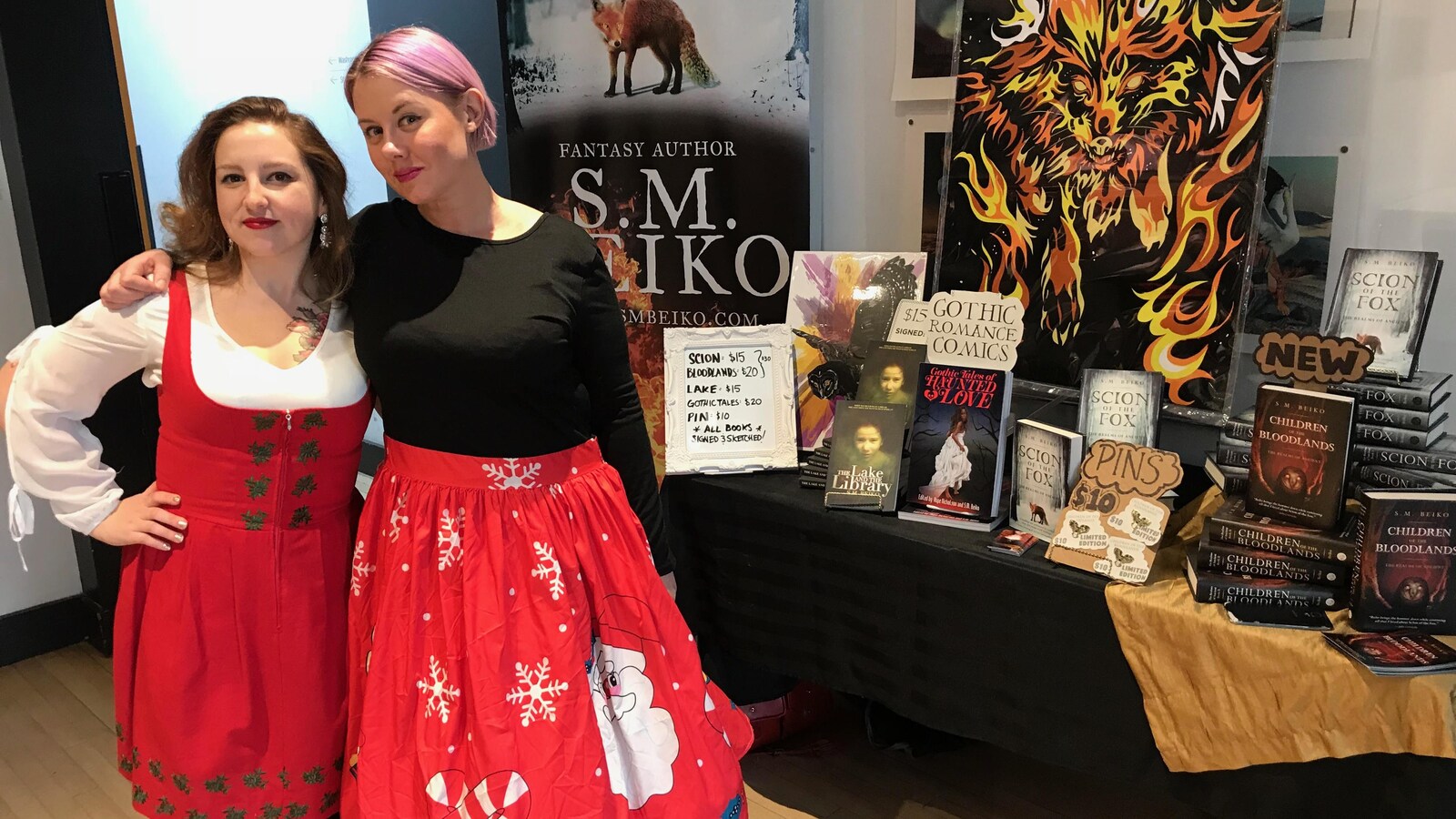 Deux femmes en robes festives se tiennent par l'épaule devant une pile de bandes dessinées et de livres de littérature fantastique.