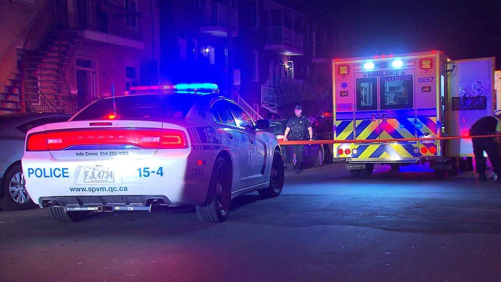Une voiture de police est stationnée dans une rue résidentielle avec ses gyrophares allumés derrière un cordon de sécurité et une ambulance qui a aussi ses gyrophares allumés dans la nuit.