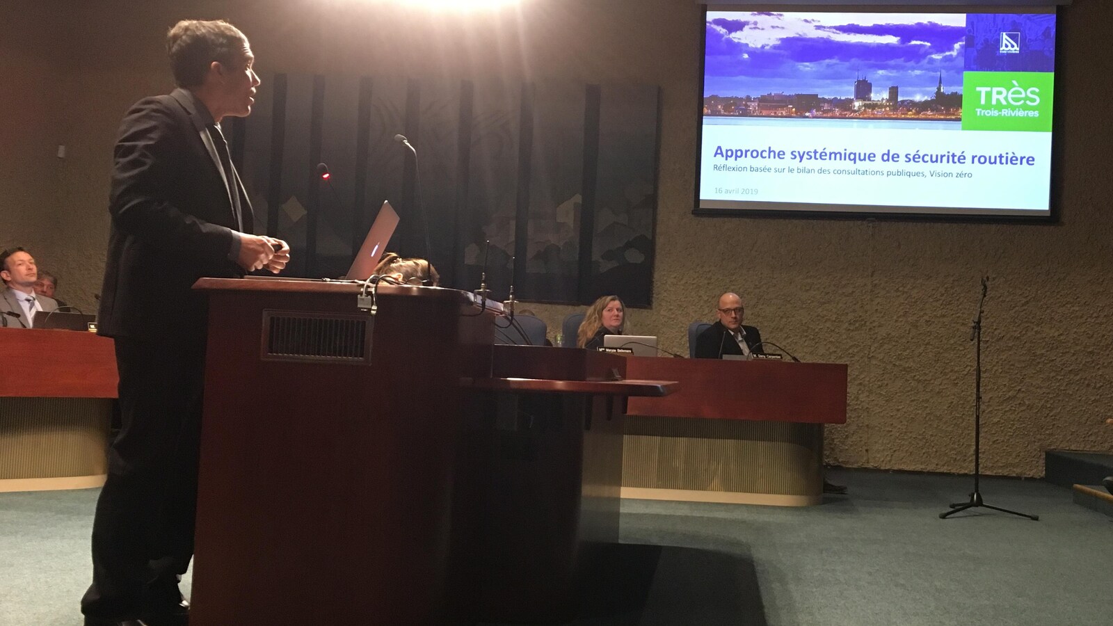 Un homme se tient derrière un bureau, il fait une présentation avec des images projetées sur une toile dans une salle de conseil municipal.