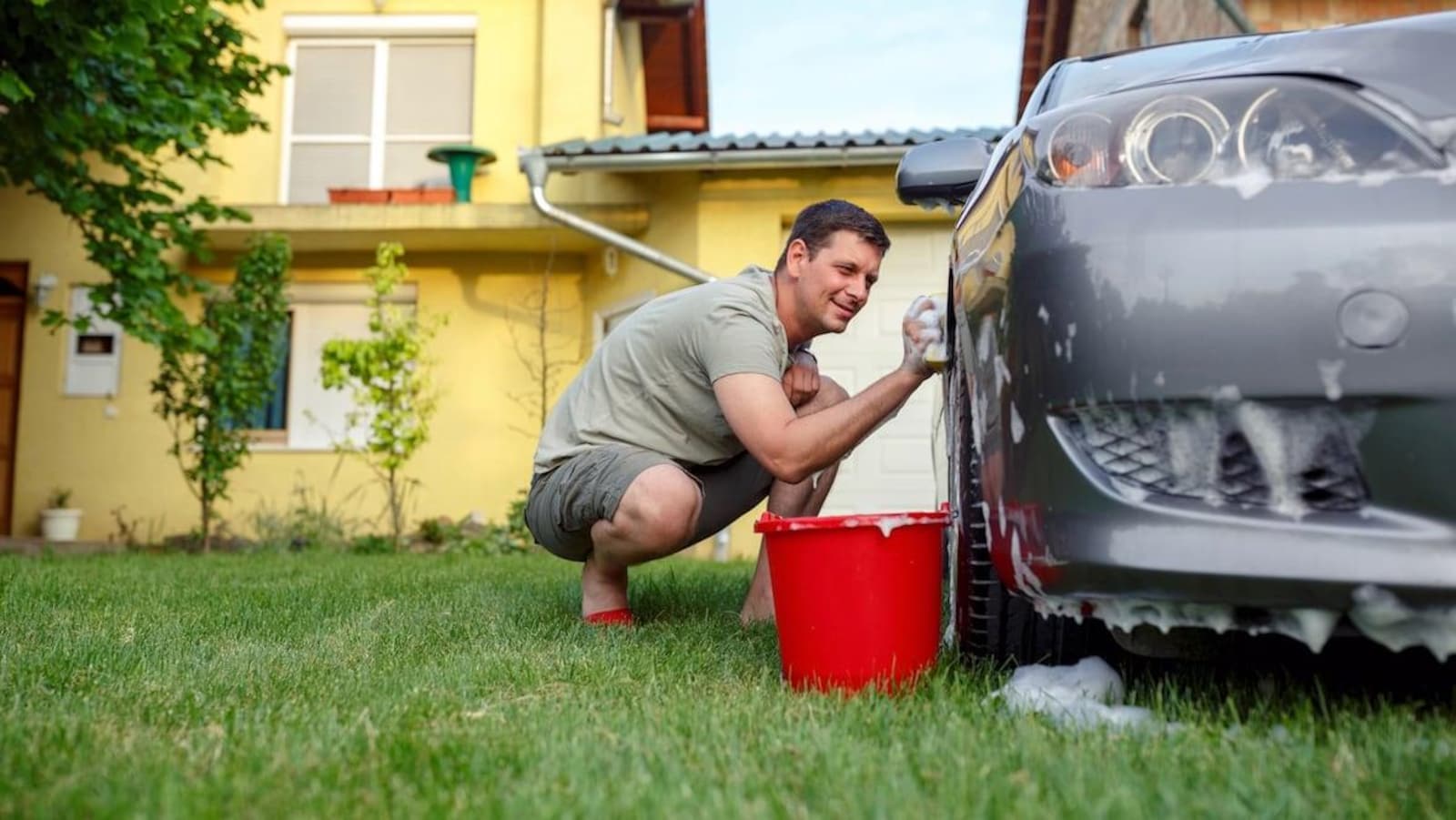 Un homme nettoie son véhicule avec une éponge, un seau d'eau et du savon.