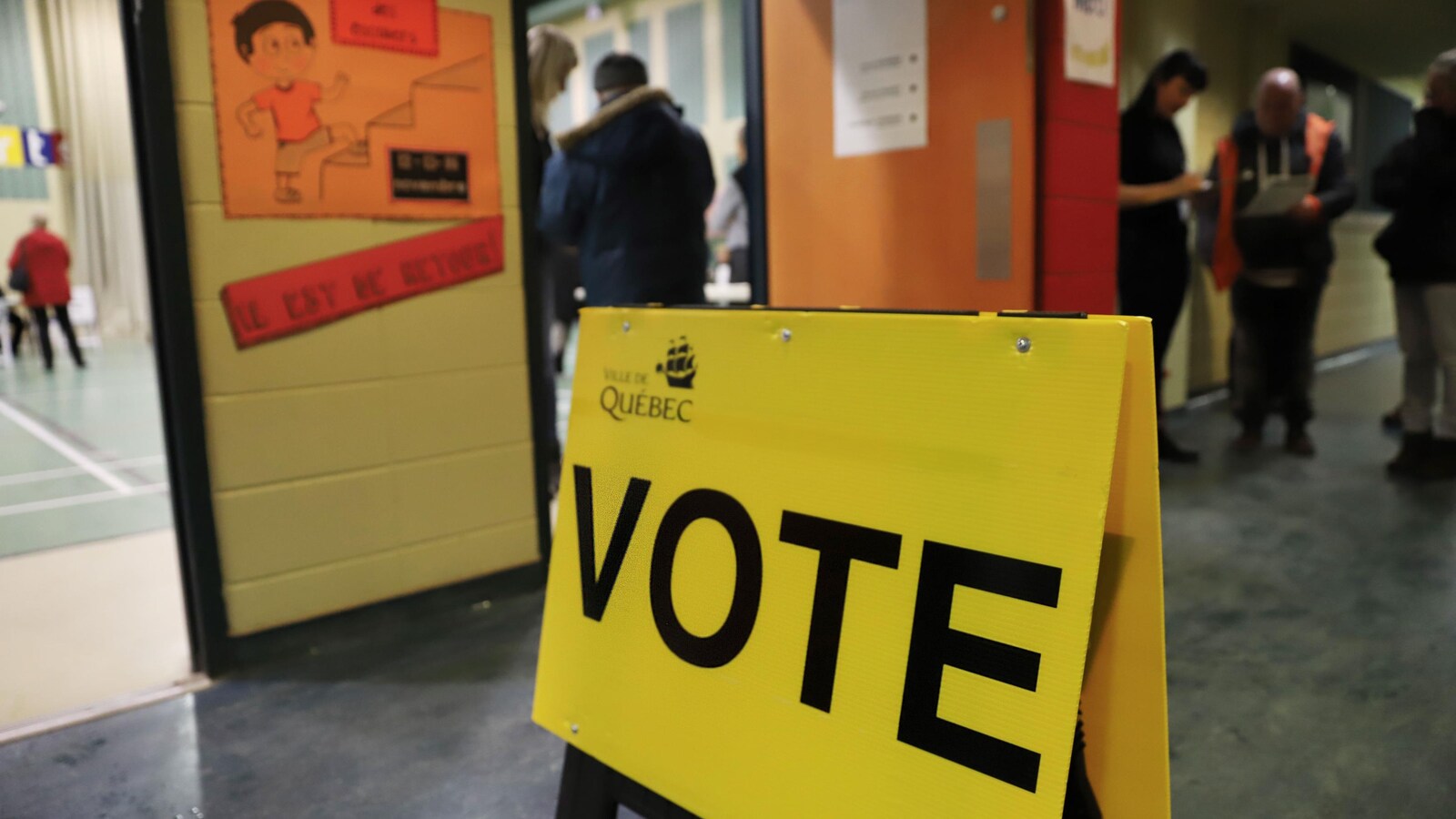 Affiche de bureau de vote installée dans le corridor d'une école avec des citoyens qui attendent leur tour pour voter.