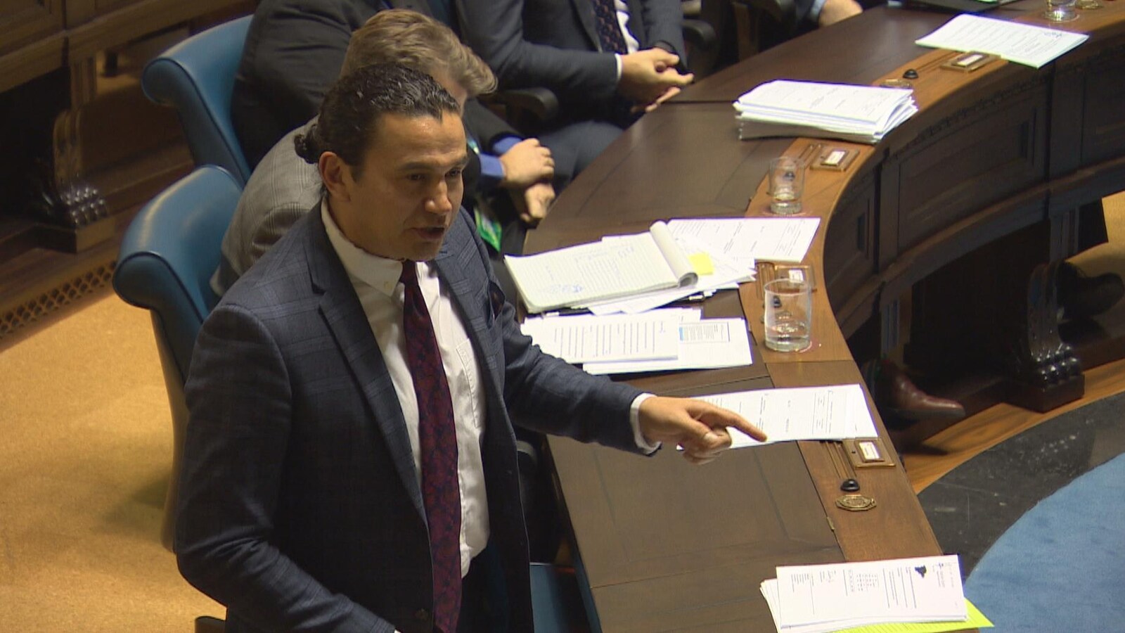 Wab Kinew debout en chambre, vu de profil, pointe du doigt pour appuyer son propos.