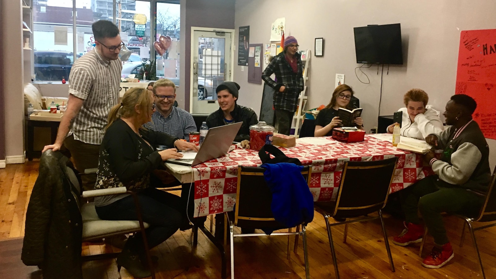 Sept personnes sont autour d'une table, l'air rieur, une autre marche vers la table, une est absorbée par sa lecture.