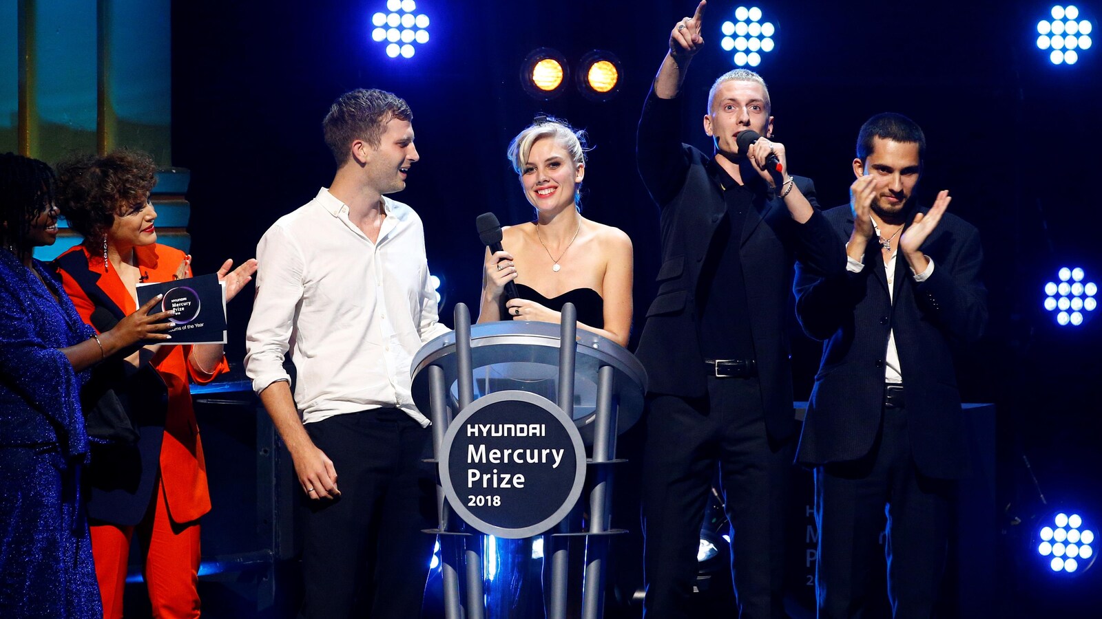 Six personnes sont sur scène dont les quatre membres du groupe qui sourient, applaudissent et lèvent la main.
