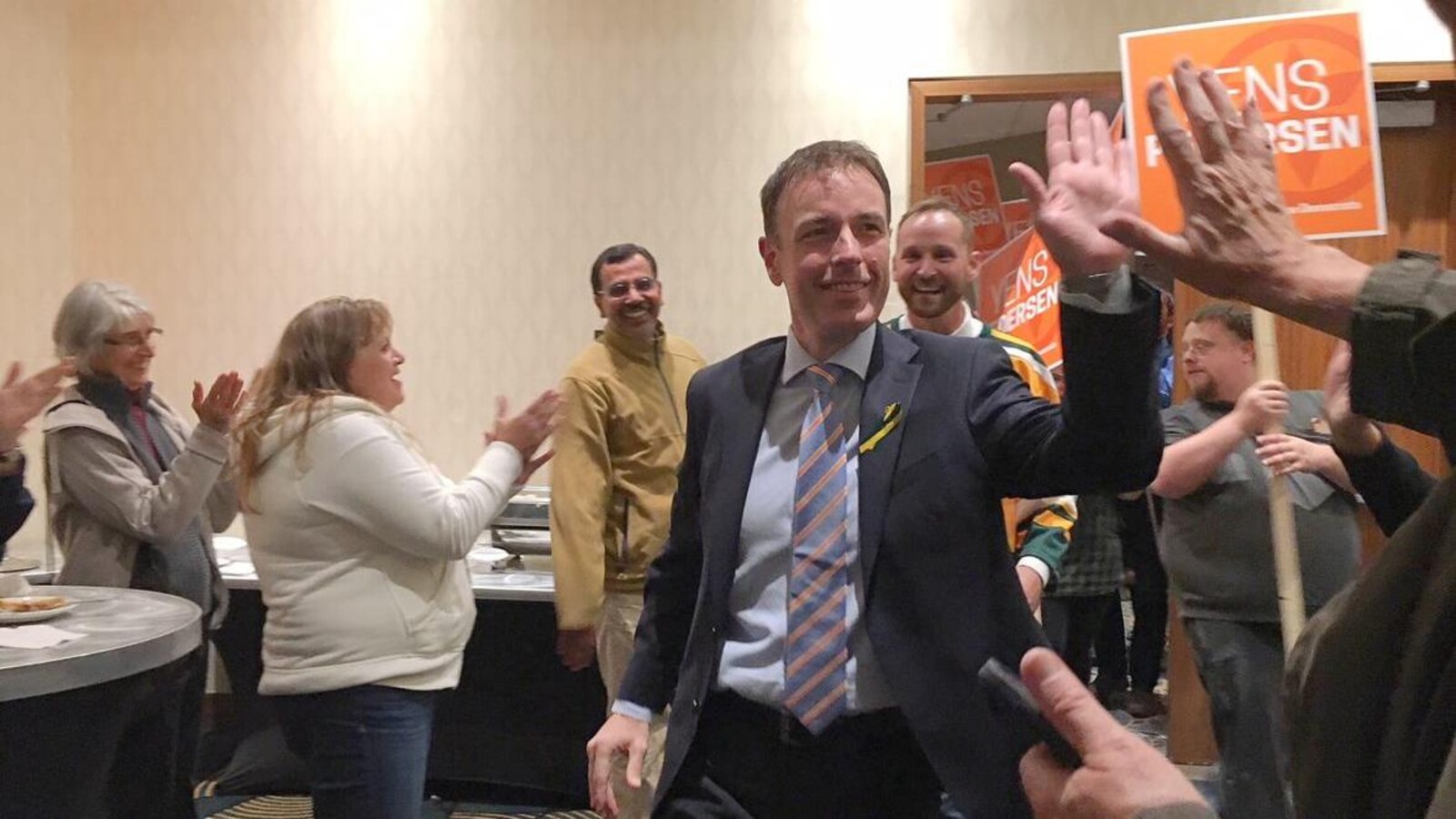 Le gagnant de l'élection tape dans la main d'un partisan. On aperçoit le chef du parti, Ryan Meili, derrière lui. Les deux sont applaudis à leur entrée dans la pièce.