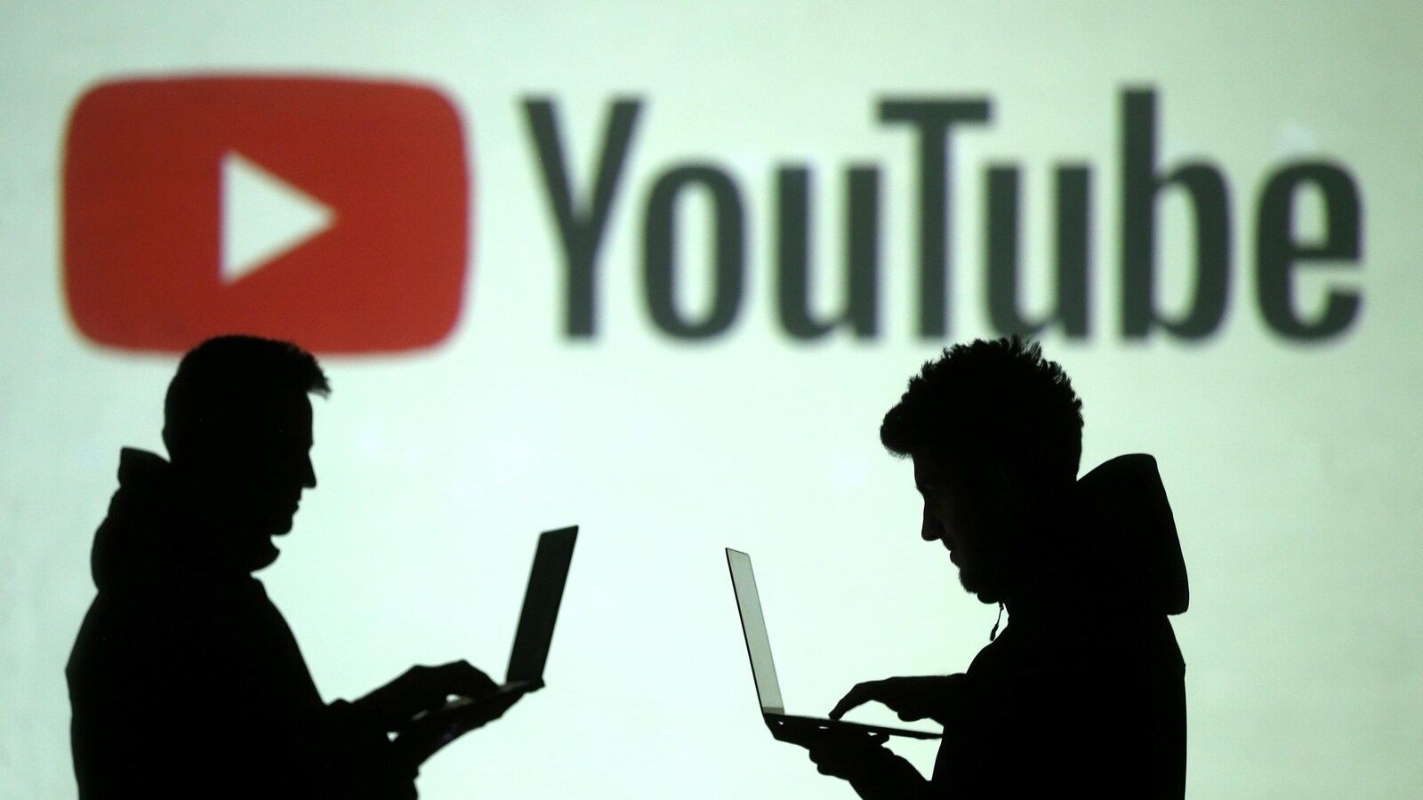 Des silhouettes de personnes utilisant des ordinateurs portables sont visibles devant un immense logo de l'entreprise YouTube.