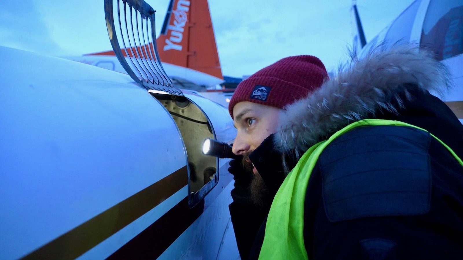 Un homme chaudement habillé, tient une lampe torche dans sa main droite. Il regarde à l'intérieur d'une pièce métallique. Derrière, il est écrit : Yukon sur ce qui semble être une aile d'avion.