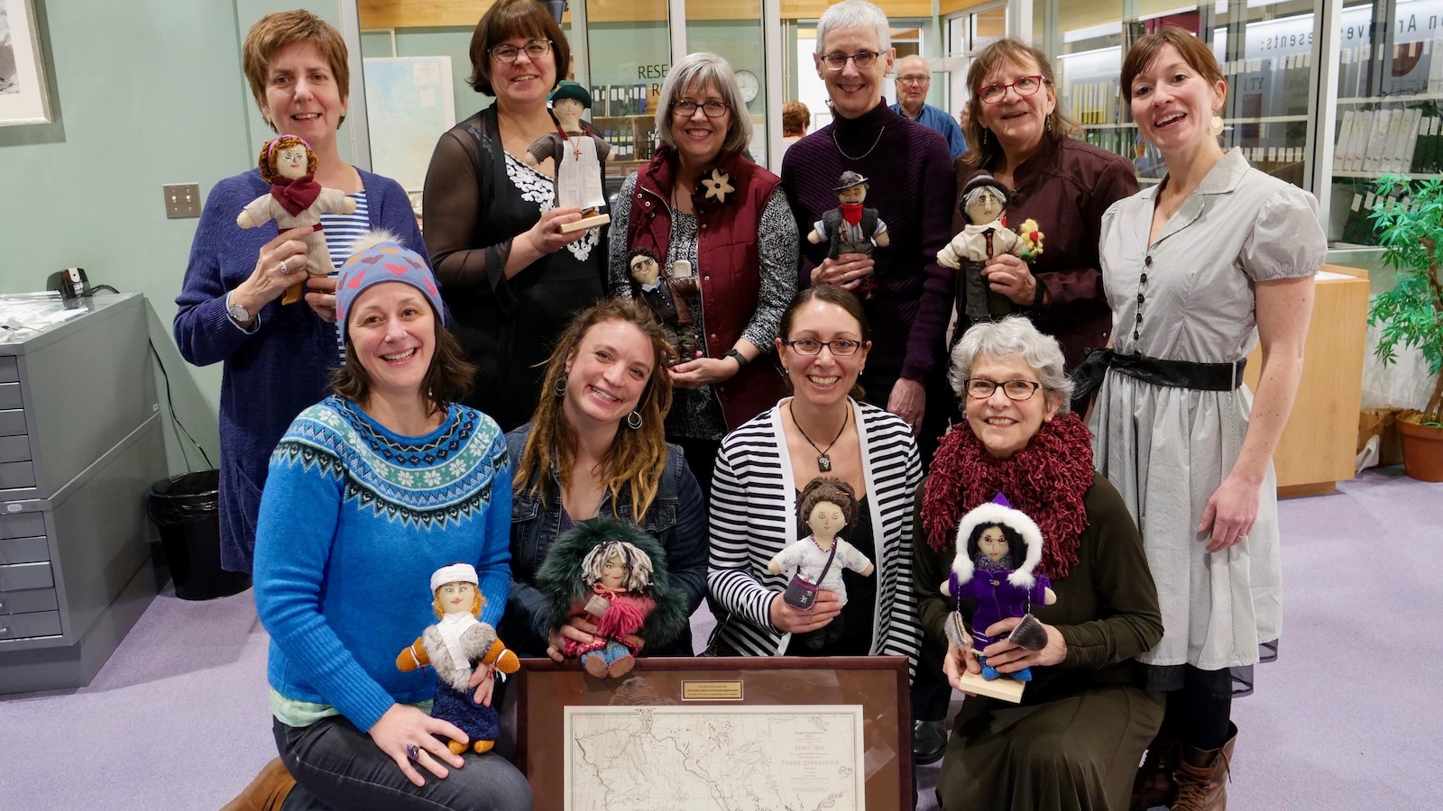 Dix femmes posent pour la photo avec des poupées en tissu dans les mains. Un cadre avec une carte géographique du Yukon est posé devant elles.
