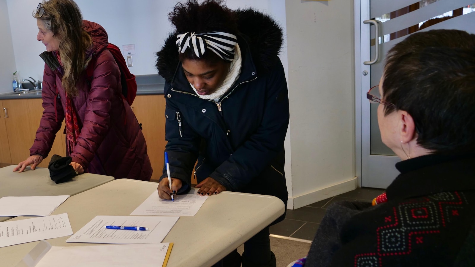 Une femme debout est en train de signer un papier. Devant elle, une dame est assise et la regarde. À côté d'elle, une autre femme est debout et semble s'adresser à une personne hors de l'image.