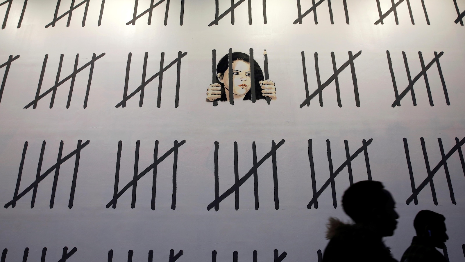 Deux passants marchent devant un mur blanc. Le visage de la journaliste et artiste Zehra Dogan y a été dessiné par Banksy derrière des barreaux, dont un est aussi un crayon. D'autres traits noirs représentent les jours passés en prison par la Turque.