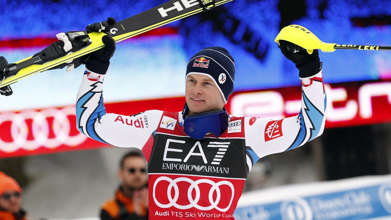 Il tient ses skis dans la main droite et un bâton dans la gauche.
