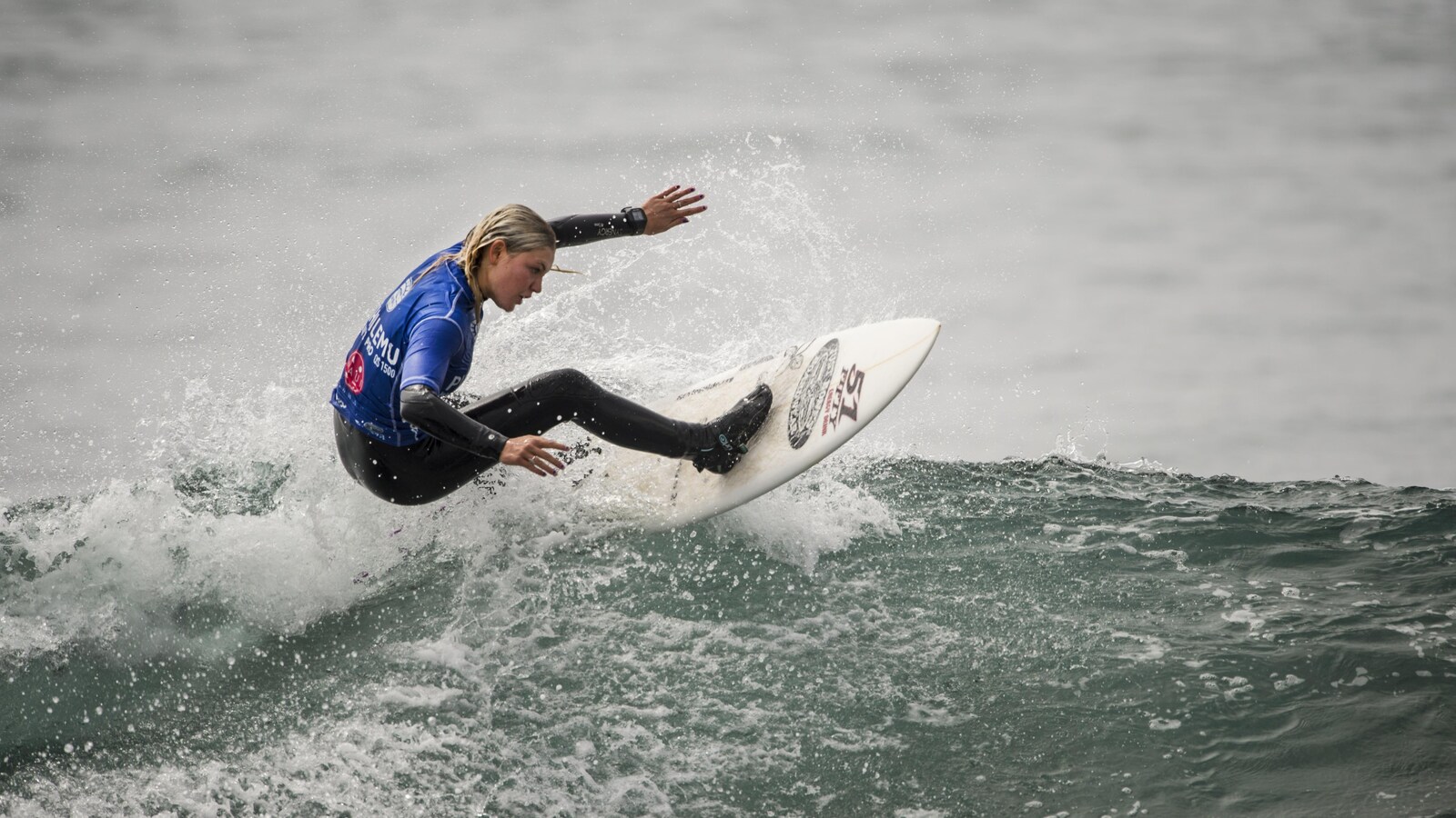 Une jeune femme blonde, dans l'eau, debout sur une planche de surf.