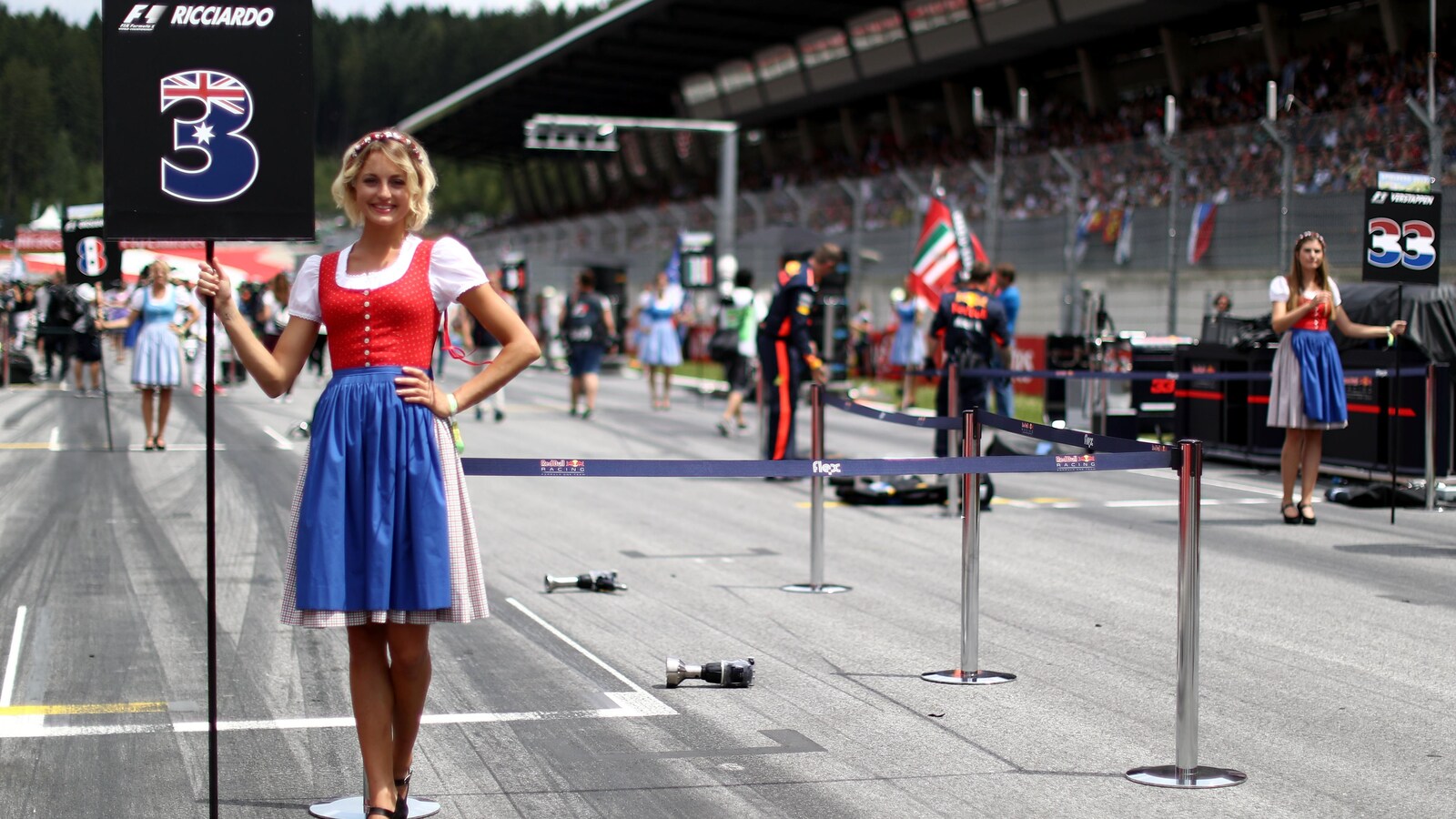 Une jolie jeune femme vêtue d'un costume traditionnel autrichien pose fièrement en tenant un panneau indiquant le nom du pilote Daniel Ricciardo et le numéro de son couloir (3).