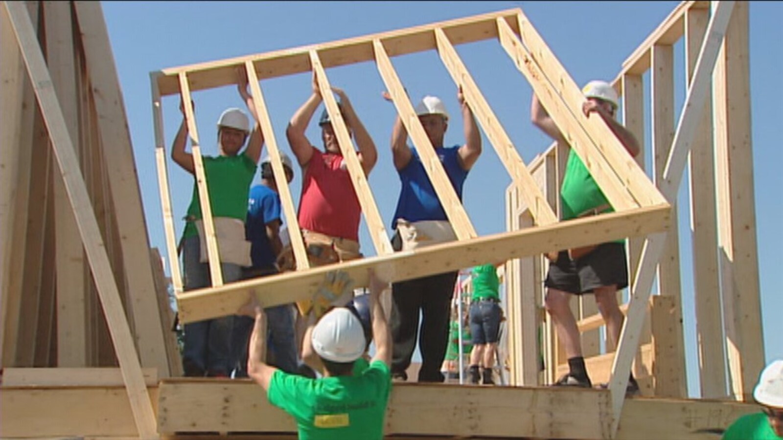 Plusieurs personnes avec des casques de protection installent la charpente d'un mur d'une maison en construction.
