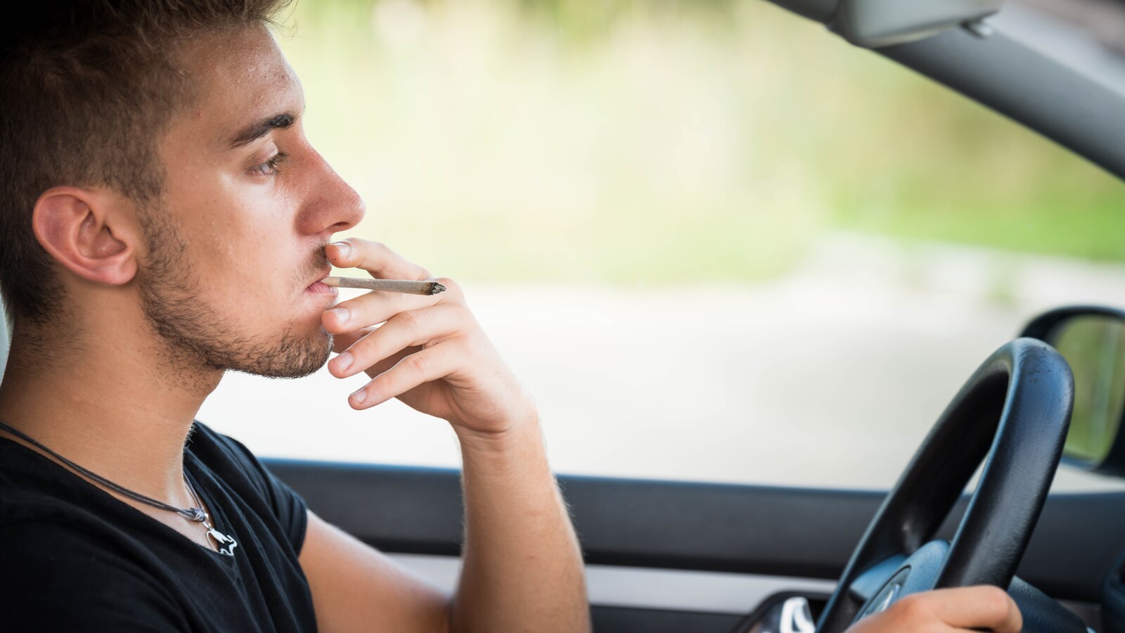 Un jeune homme conduit une voiture en fumant son joint de cannabis.