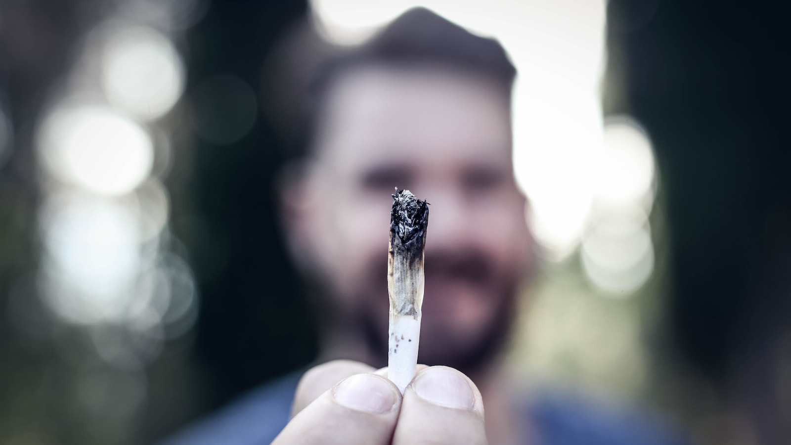 Un homme tient un joint de cannabis entre ses doigts et il le montre devant de lui.