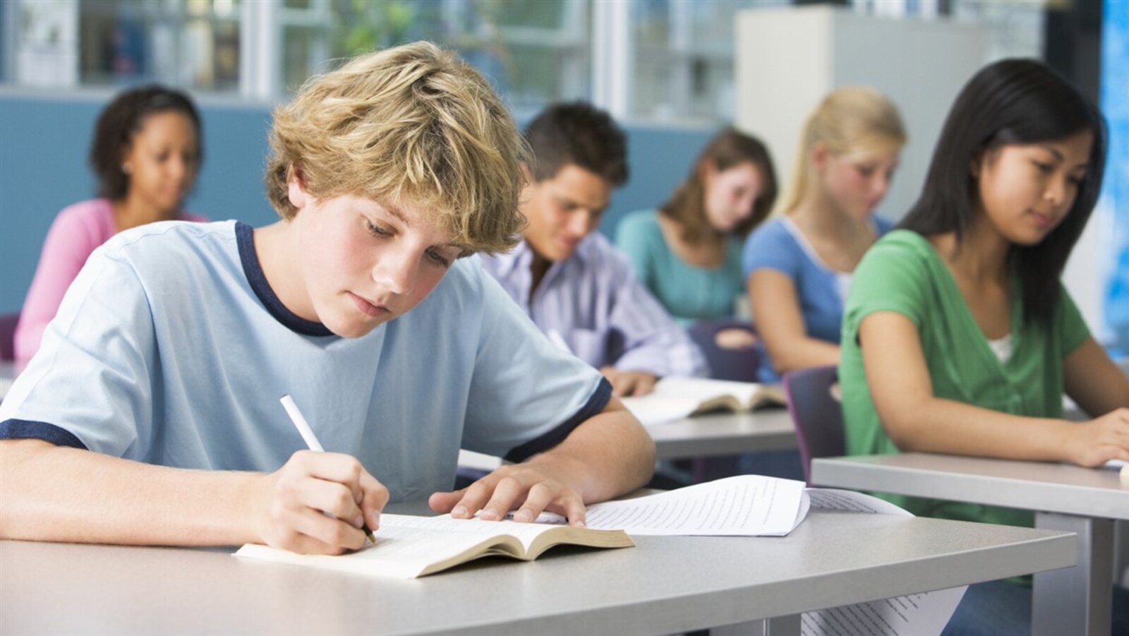 Des élèves du secondaire assis en classe. Au premier plan, un jeune lit et prend des notes.