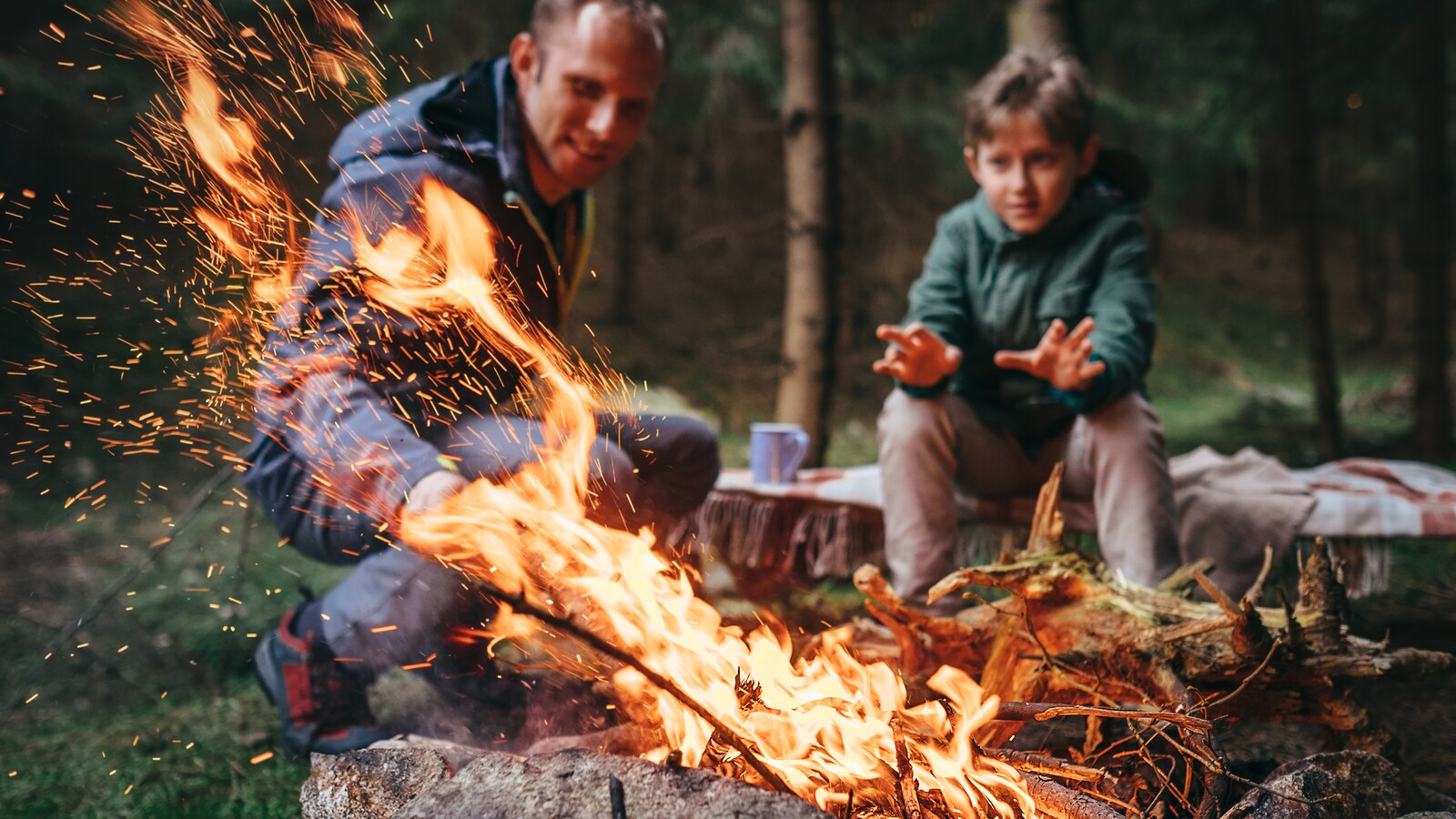 Un père et son fils devant un feu de camp. Le jeune se réchauffe les mains pendant que son père alimente le brasier.