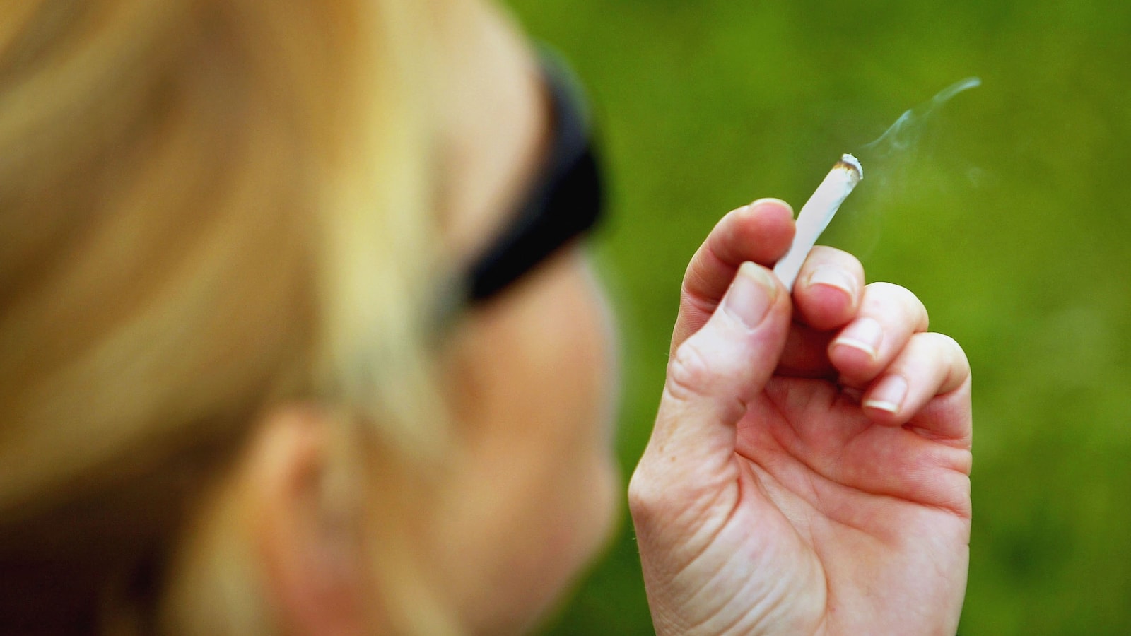 Une personne portant des lunettes de soleil tient un joint dans sa main gauche. De la fumée s'en échappe.