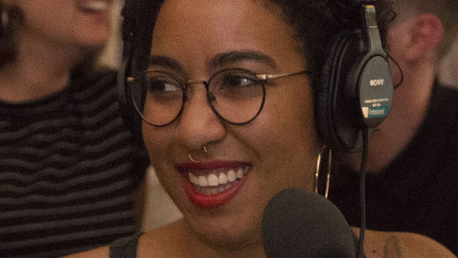 Marilou Craft en gros plan de trois quart, devant un micro, un casque sur la tête, souriant. Elle porte un anneau dans le nez.