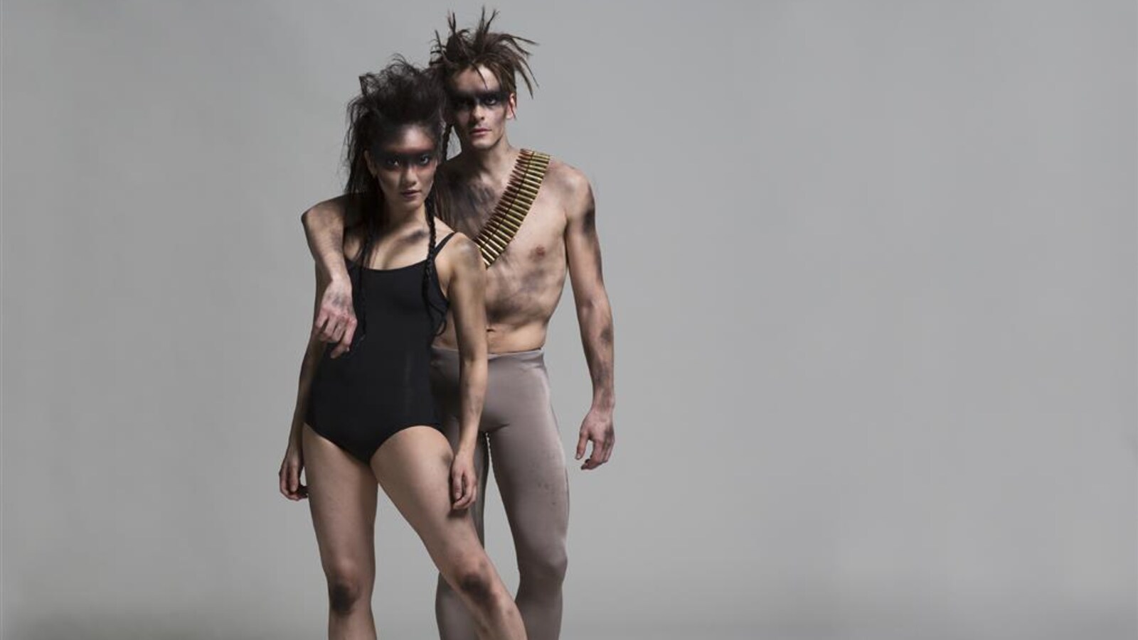 Une danseuse et un danseur sont debout sur une scène dénudée avec un fond gris. L'homme pose un bras sur l'épaule de la femme. Ils sont tous deux sales et leurs chevelures sont ébouriffées.