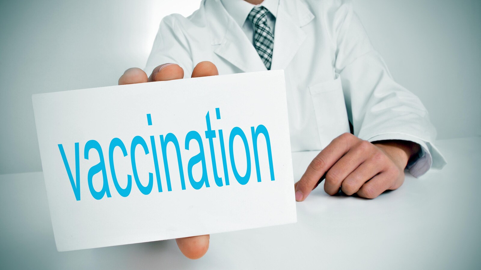 Un homme dont on ne voit pas le visage, vêtu d'une blouse blanche, tient un carton dans sa main droite où est écrit en bleu le mot vaccination.