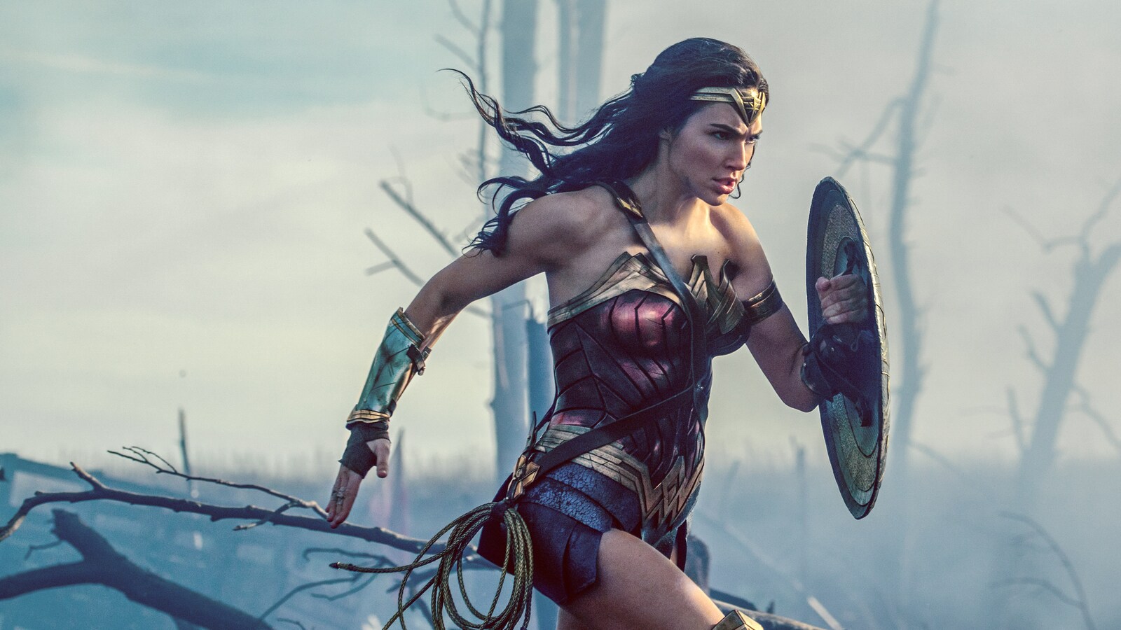 L'actrice Gal Gadot dans le costume de Wonder Woman, court dans une forêt enfumée, bouclier sur la bras gauche.
