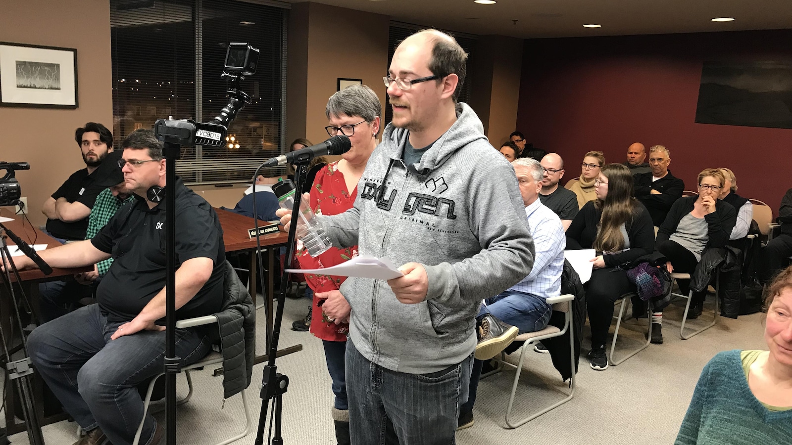 Un homme avec un chandail gris parle au micro devant des spectateurs au conseil municipal.