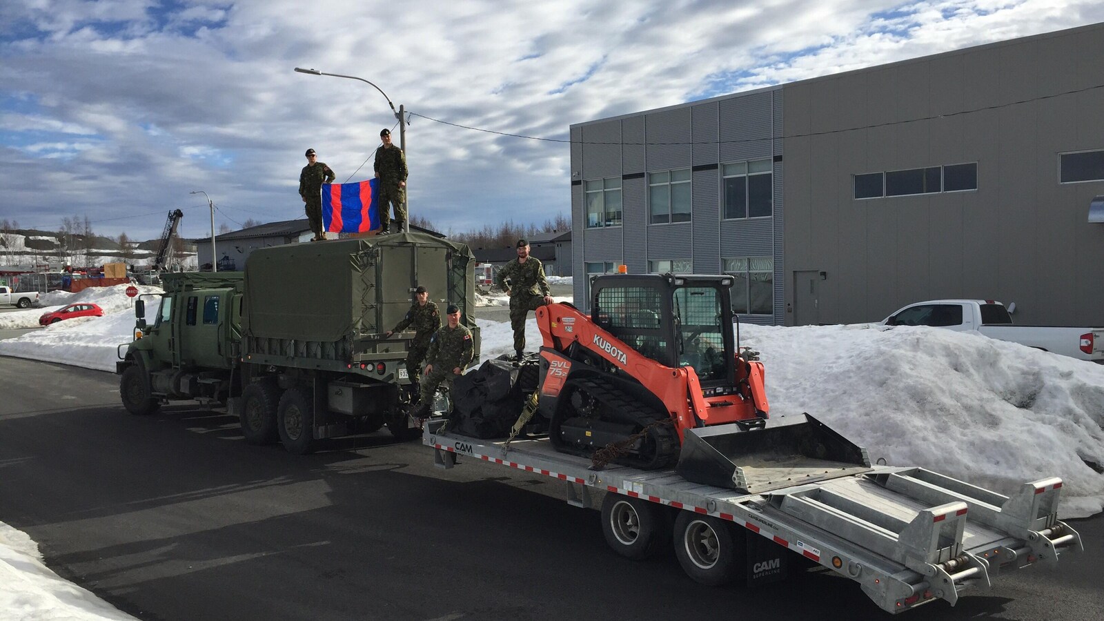 Des soldats debout sur un camion vert de l'armée et sur un bobcat dans une remorque prennent une photo avec le drapeau du Génie Royale Canadien.