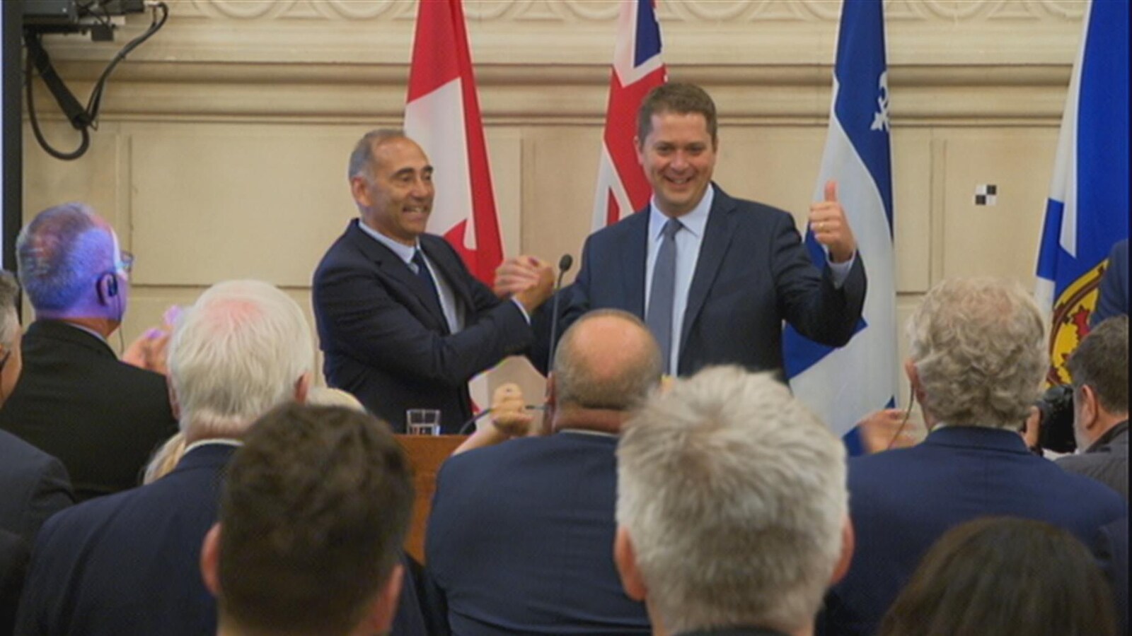 Richard Martel serre la main d'Andrew Sheer qui tient un pouce en l'air d'un air triomphal sur une tribune devant les députés qui font une ovation.