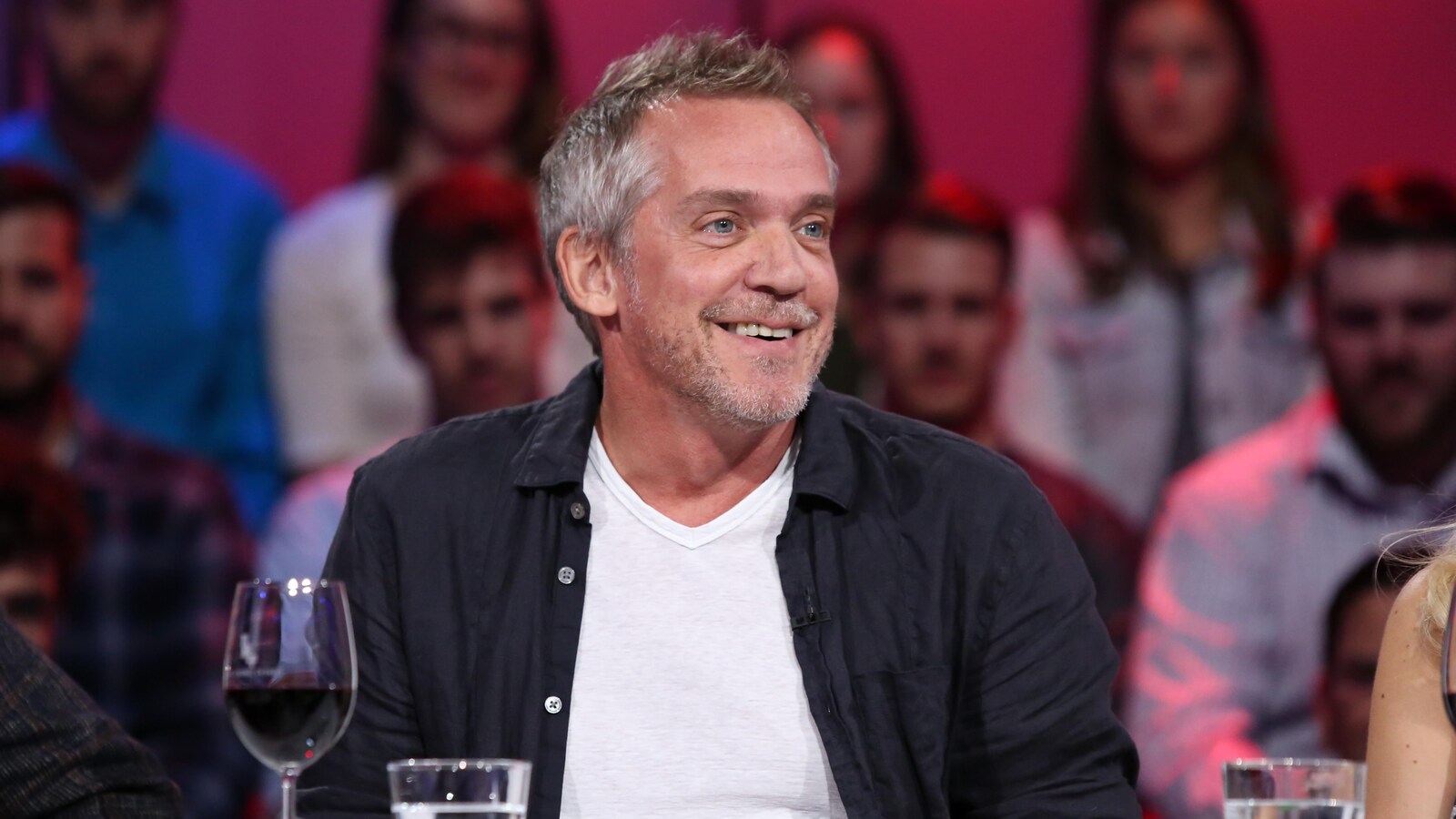 Il sourit. Il porte un chandail blanc et une chemise sombre.