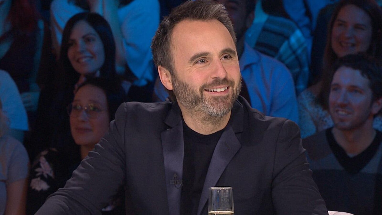 Il sourit. Il porte un chandail et un veston bleu foncé.